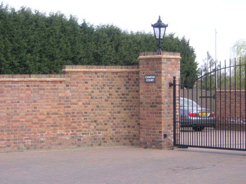 Stepped brick boundary wall and gate pillar with a lantern beside black metal gates